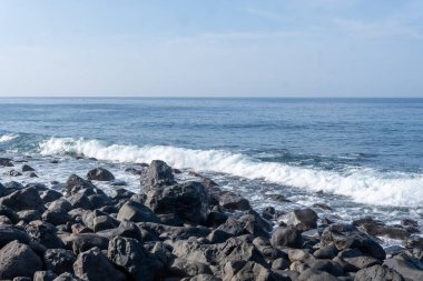 Panoramic view of a rugged coastline in Flores featuring dark volcanic rocks, white crashing waves, and a clear blue horizon.