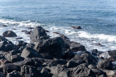 Endonezya 'nın Flores kentindeki engebeli bir sahilde büyük siyah volkanik taşlara çarpan mavi deniz dalgalarının güçlü bir sahnesi..