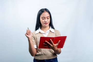 Endonezyalı genç bir kadın, elinde kalem tutarken özenle kırmızı defterine bakıyor. Yaratıcı kullanım için beyaz bir arka planda izole edilmiş..