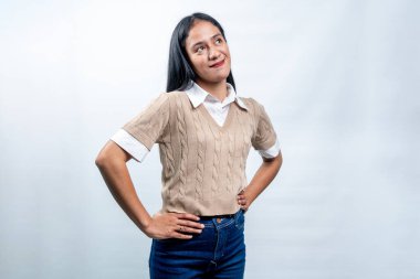 Endonezyalı genç bir kadın kollarıyla kendine güvenerek ayakta duruyor, gülümseyerek yukarı bakıyor. Temiz beyaz bir arkaplanda pozitif ve iddialı bir ifade.