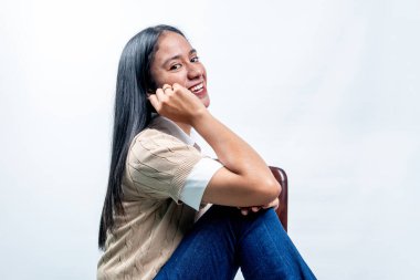 Neşeli genç Endonezyalı kadın oturuyor ve gülümsüyor, çenesini elinde dinlendiriyor. Dost canlısı ifade, şık günlük tarz, beyaz üzerine izole edilmiş..