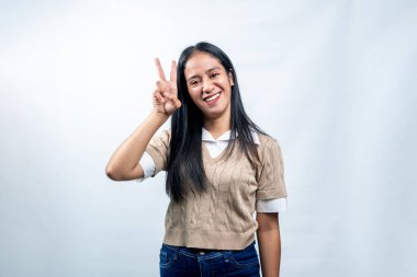 Mutlu genç Güneydoğu Asyalı kadın kameraya gülümsüyor ve temiz beyaz bir arkaplana karşı zafer veya barış işareti gösteriyor..