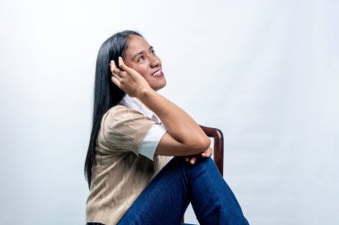 Neşeli genç bir Güneydoğu Asyalı kadın büyük bir gülümsemeyle, yan görünüşüyle temiz beyaz bir arka planda bir sandalyede oturuyor..