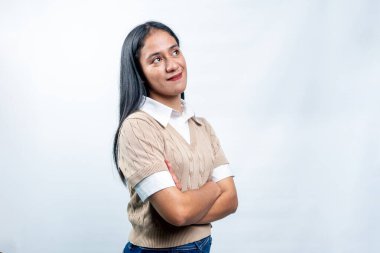Başarılı genç Güneydoğu Asyalı kadın, günlük iş kıyafetleri içinde kolları bağlı oturuyor, beyaz bir arka planda kendinden emin bir gülümsemeyle başka tarafa bakıyor..