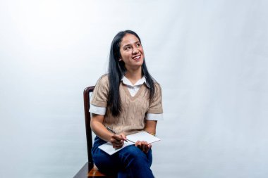 Mutlu Güneydoğu Asyalı kadın, beyaz bir arka planda elinde kalem ve defter tutarken ilham verici bir ifadeyle yukarı bakıyor..