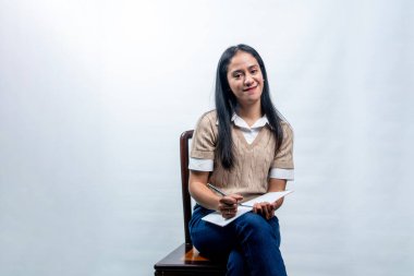 Tahta bir sandalyede oturan, elinde temiz beyaz bir arka planda defter ve kalem tutan, kendine güvenen Güneydoğu Asyalı bir kadın..