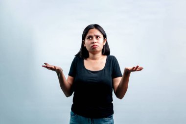 Kafası karışık ve şüpheli bir ifadeyle yukarı bakan genç Asyalı kadın, beyaz bir stüdyonun arka planında dururken ellerini silkiyor..