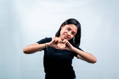 Siyah tişörtlü neşeli Endonezyalı kadın parmaklarıyla bir kalp sembolü oluşturuyor ve gülümsüyor, beyaz arka planda izole edilmiş..