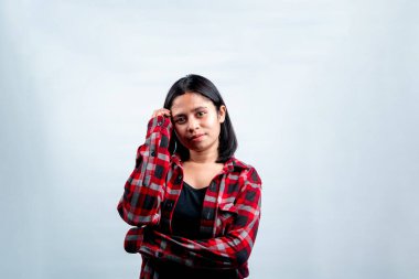 Genç bir Güneydoğu Asyalı kadın kafasına dokunurken sakince gülümsüyor, siyah bir tişörtün üzerine kırmızı bir flanel giyiyor..