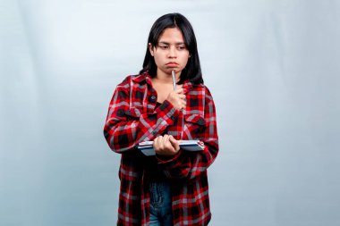 Kırmızı ekose gömlekli düşünceli genç bir Güneydoğu Asyalı kadın, elinde düz bir arkaplana karşı bir defter tutarken çenesine kalem dayıyor..