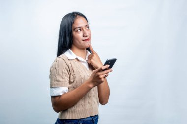 Süveter giyen, elinde telefon tutan ve çenesine dokunan Güneydoğu Asyalı düşünceli genç bir kadın. Beyaz arkaplanda fotokopi alanı olan stüdyo portresi.