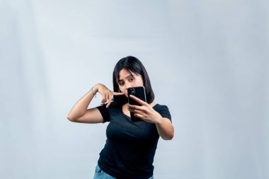 Siyah tişörtlü neşeli genç bir kadın selfie çekerken ya da akıllı telefonuyla beyaz bir arkaplana karşı video kaydederken neşeli poz veriyor..