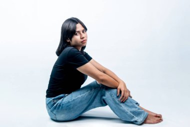 Siyah gömlekli ve yırtık kotlu genç bir kadın beyaz bir zeminde yan profil pozunda oturuyor ve sakin bir ifadeyle kameraya bakıyor..