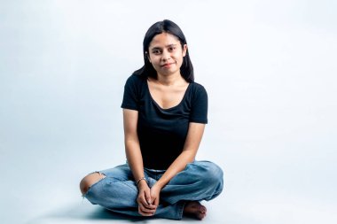 Mutlu genç bir kadının portresi, yerde rahat bir şekilde oturuyor, yumuşak bir gülümsemeyle, günlük siyah tişört giyiyor ve beyaz arka plana karşı kot pantolon giyiyor..
