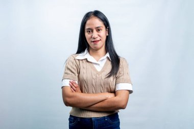 Kolları çapraz duran bej rengi yelek ve beyaz gömlekli genç bir Asyalı kadın beyaz arka planda sakin bir ifadeyle kameraya bakıyor..