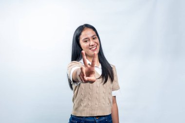 Beyaz tişörtlü, bej yelekli mutlu bir Asyalı kadın beyaz bir stüdyo arka planında, parmaklarıyla V işareti ya da barış hareketi yapıyor..