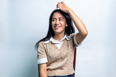 Beyaz tişörtlü, bej rengi yelekli, kafası karışık ama mutlu bir ifadeyle başını beyaz arka planda tutan genç bir Asyalı kadın..