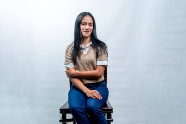 Beyaz gömlekli, bej renkli bir kadın tahta bir sandalyede oturuyor. Nazikçe gülümsüyor ve elleri çapraz, beyaz tenli..