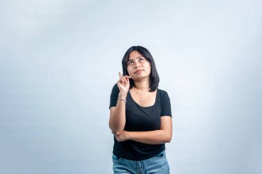 Siyah tişörtlü genç bir Asyalı kadın dikkatlice yukarı bakıyor ve beyaz bir arka planda yeni bir fikri varmış gibi bir parmağını yukarı kaldırıyor..