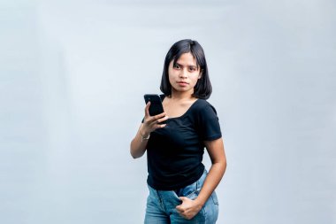Siyah tişörtlü genç bir Asyalı kadın, beyaz arka planda akıllı telefonunu tutarken ciddi ve kendinden emin bir ifadeyle ayakta duruyor..