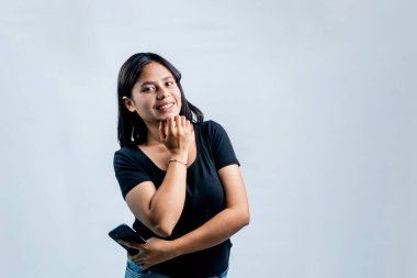 Cep telefonu tutan mutlu, genç bir Asyalı kadın, temiz beyaz bir arkaplanda eli çenesinde neşeli bir şekilde gülümsüyor..