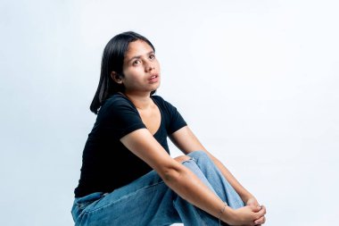 Siyah gömlekli ve kotlu genç bir Asyalı kadın beyaz bir zeminde oturuyor, umut dolu, hayalperest ya da ruhani bir ifadeyle yukarı bakıyor..