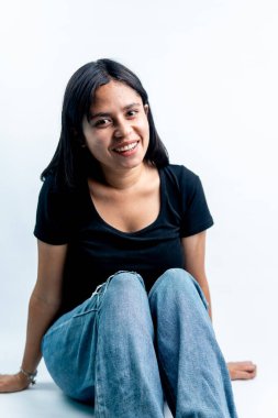 Beyaz arka planda oturan neşeli Asyalı kız kameraya bakıyor. Stüdyo aydınlatmasında mutlu bir genç kadının sıradan bir portresi..