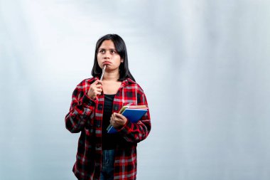 Kırmızı ekose gömlekli, elinde kitaplarla çenesine kalem tutarak ilham arayan düşünceli genç bir kadın. Beyaz arka planda izole edilmiş stüdyo çekimi.