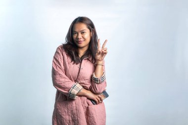 V-işareti ya da barış işareti ile poz veren pembe örtülü bir ceket giyen mutlu Endonezyalı kadın. Yaşam tarzı temaları için beyaz arkaplanda izole edilmiş.