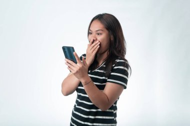Siyah çizgili tişörtlü Endonezyalı genç bir kadın şok olmuş görünüyor ve telefonu tutarken ağzını kapatıyor. İnançsızlık mı, kötü haber mi?.