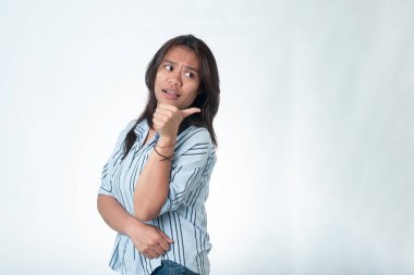 Portrait of an Indonesian woman in a blue striped shirt pointing her thumb over her shoulder. She has a curious or doubtful expression, looking to the side. Isolated on white.