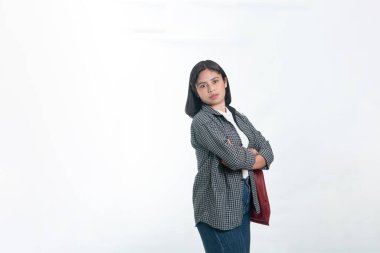 Ekose gömlekli ve kot pantolonlu Endonezyalı kadın elinde kırmızı bir çantayla dikiliyor. Kolları bağlı, kendinden emin bir ifade. Beyaz arkaplanda izole.