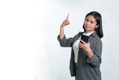 Endonezyalı genç bir kadının portresi, kısa siyah saçlı, kareli fanila gömlekli, akıllı bir telefon tutan ve işaret parmağını yukarı doğru gösteren. Beyaz arkaplanda izole.