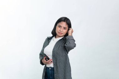 Kısa saçlı Endonezyalı bir kadının portresi, kareli bir fanila gömlek giyiyor, bir elinde akıllı bir telefon tutarken diğer eliyle zarif bir şekilde saçına dokunuyor. Beyazda izole edilmiş.
