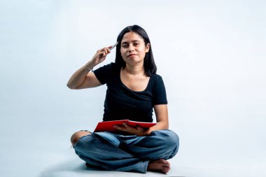 Siyah tişörtlü Endonezyalı kadın çapraz bacaklı kırmızı kitap ve kalemle kafa kafaya oturuyor. Üniversite eğitimi kavramı, beyin fırtınası ve beyaz arka planda izole edilmiş yaratıcı düşünce.