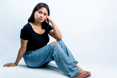 Siyah tişörtlü ve yırtık kotlu gülümseyen Endonezyalı kadın baş aşağı yerde oturuyor. Sıkıntı kavramı, beklemek ve yorgun gençlik yaşam tarzı beyaz arka planda izole edilmiş..