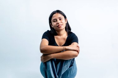 Beyaz bir arka plan karşısında oturan ve dizlerini kucaklayan Endonezyalı genç bir kadının portresi..