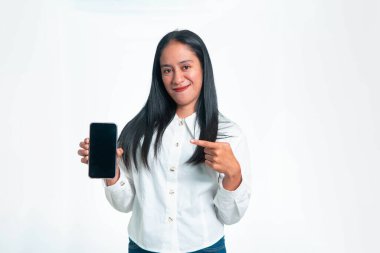 Beyaz gömlekli neşeli bir Endonezyalı kadın beyaz bir stüdyonun arka planına işaret ederken boş ekranlı bir akıllı telefon sergiliyor..