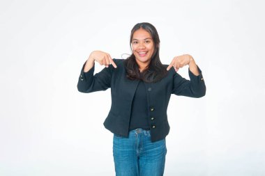 Siyah desenli ceket ve kot pantolon giyen gülümseyen Endonezyalı kadının portresi, kameraya heyecanlı bir ifadeyle bakarken her iki işaret parmağını da aşağı doğru, beyaz arka planda izole bir şekilde gösteriyor..