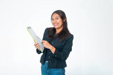 Portrait of Indonesian woman wearing black textured jacket and blue jeans, holding green clipboard and pointing at it with pen while smiling at camera, isolated on white background.