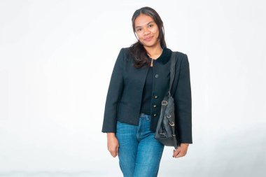 Siyah ceket ve kot pantolon giyen, siyah omuz çantası taşıyan, beyaz arka planda poz veren gülümseyen Endonezyalı kadın portresi..