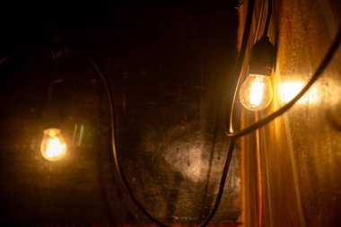 Macro shot of a single warm, glowing vintage filament light bulb from a string, with another blurred bulb creating bokeh in the dark background.