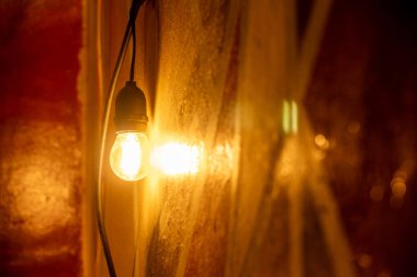 A single hanging vintage light bulb glowing with warm orange light, casting a strong reflection and cozy ambiance against a textured golden-brown wall.