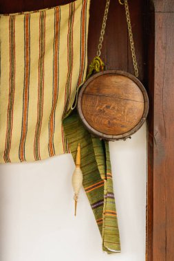 Ahşap bir duvarda asılı el yapımı ahşap bir matara ve renkli dokuma tekstil kompozisyonu. Sıcak tonlar ve geleneksel dokular kırsal mirası ve zanaatkarlığı çağrıştırıyor..
