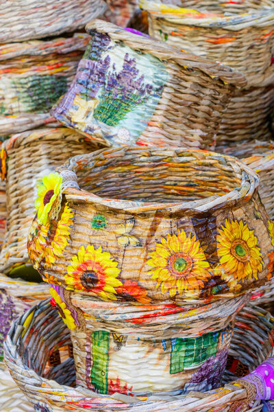 Stacked woven baskets adorned with sunflower and lavender motifs are displayed under sunlight at a craft market. The detailed textures and vibrant colors reflect creativity, craftsmanship, and rustic charm.