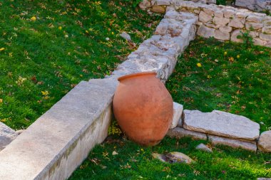 Yeşil çimenler ve dağınık sonbahar yapraklarıyla çevrili eski taş duvarların yanında büyük bir terracotta amfora yatar. Güneş ışığı, huzurlu bir bahçe sahnesinde tarihi ve doğayı harmanlayan ılık toprak tonlarını güçlendirir..