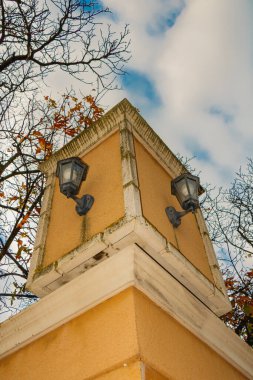Her iki yanına da iki klasik siyah duvar lambası monte edilmiş sarı bir bina sütunu. Sahne, mimarinin dokusunu, çıplak ağaç dallarını ve parçalı bulutlu gökyüzünü yakalıyor. Şehir detaylarını klasik bir cazibeyle harmanlıyor..