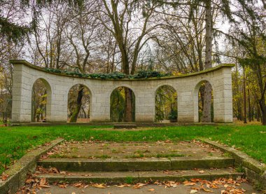 Parkta birden fazla kemerli taş bir yapı duruyor. Çevreleyen ağaçların sarı ve kahverengi yaprakları vardır. Yer düşen yapraklarla kaplı. İnsanlar yakınlarda yürürken görülebilir..