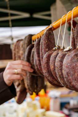 Açık hava pazarında sıra sıra sallanan sosisler arasından seçim yapan birinin elini kapatıyoruz. Görüntü, sosislerin orijinal el yapımı dokusunu, canlı arka plan renklerini ve geleneksel yerel gıda ticaretinin atmosferini vurguluyor..