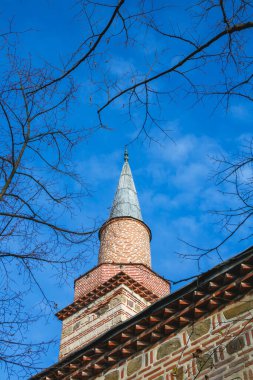 Uzun bir cami kulesi parlak mavi bir gökyüzüne karşı duruyor. Çıplak ağaç dalları, mimarisi ile bilinen tarihi bir yerde güneşli bir günde kulenin tepesini çerçeveler..